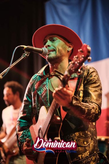 Sandro Reyes canta con guitarra, vistiendo chaqueta brillante y sombrero.