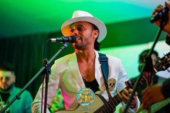 Sandro Reyes con guitarra y sombrero blanco, canta en escenario iluminado.