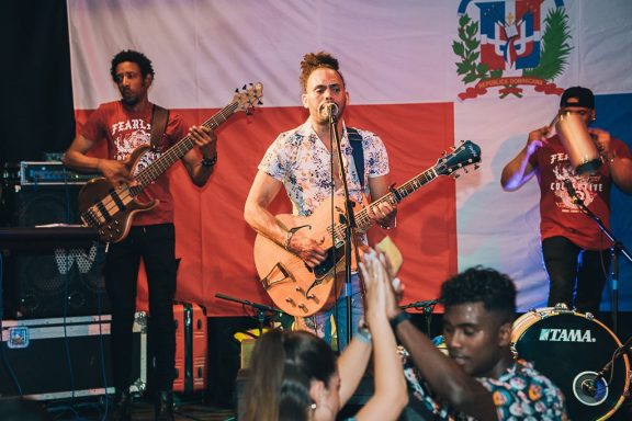 Banda de Sandro Reyes tocando en vivo frente a una bandera dominicana, con público bailando.