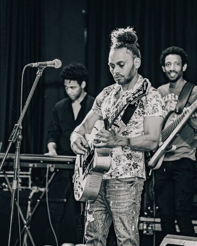 Músicos de Sandro Reyes en un escenario, uno toca la guitarra y otros dos lo acompañan.