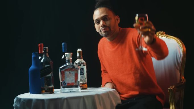 Sandro Reyes sonriente con una copa en mano, junto a varias botellas de licor en una mesa.